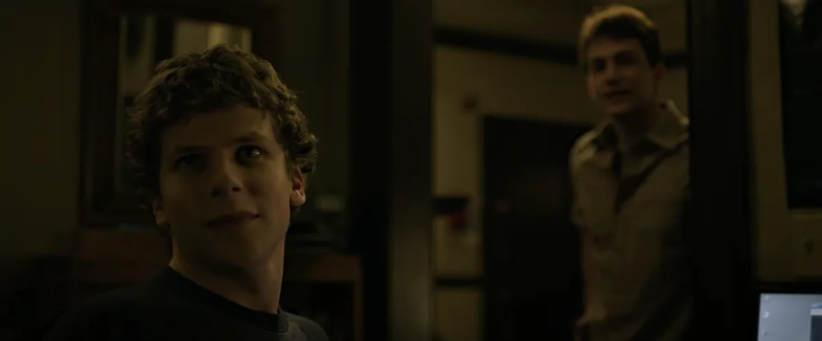 Jesse Eisenberg as Mark Zuckerberg, smiling at his monitor in a dark dorm room while Eduardo stands in the doorway behind him